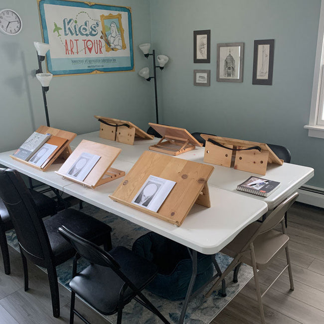A classroom or studio room with a white folding table, black chairs, and multiple wooden artist easels holding sketchbooks and prints. The room has light blue-gray walls, framed artwork, a clock, and a sign reading 'Kids Art Tour'.