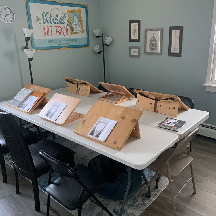 A classroom or studio room with a white folding table, black chairs, and multiple wooden artist easels holding sketchbooks and prints. The room has light blue-gray walls, framed artwork, a clock, and a sign reading 'Kids Art Tour'.